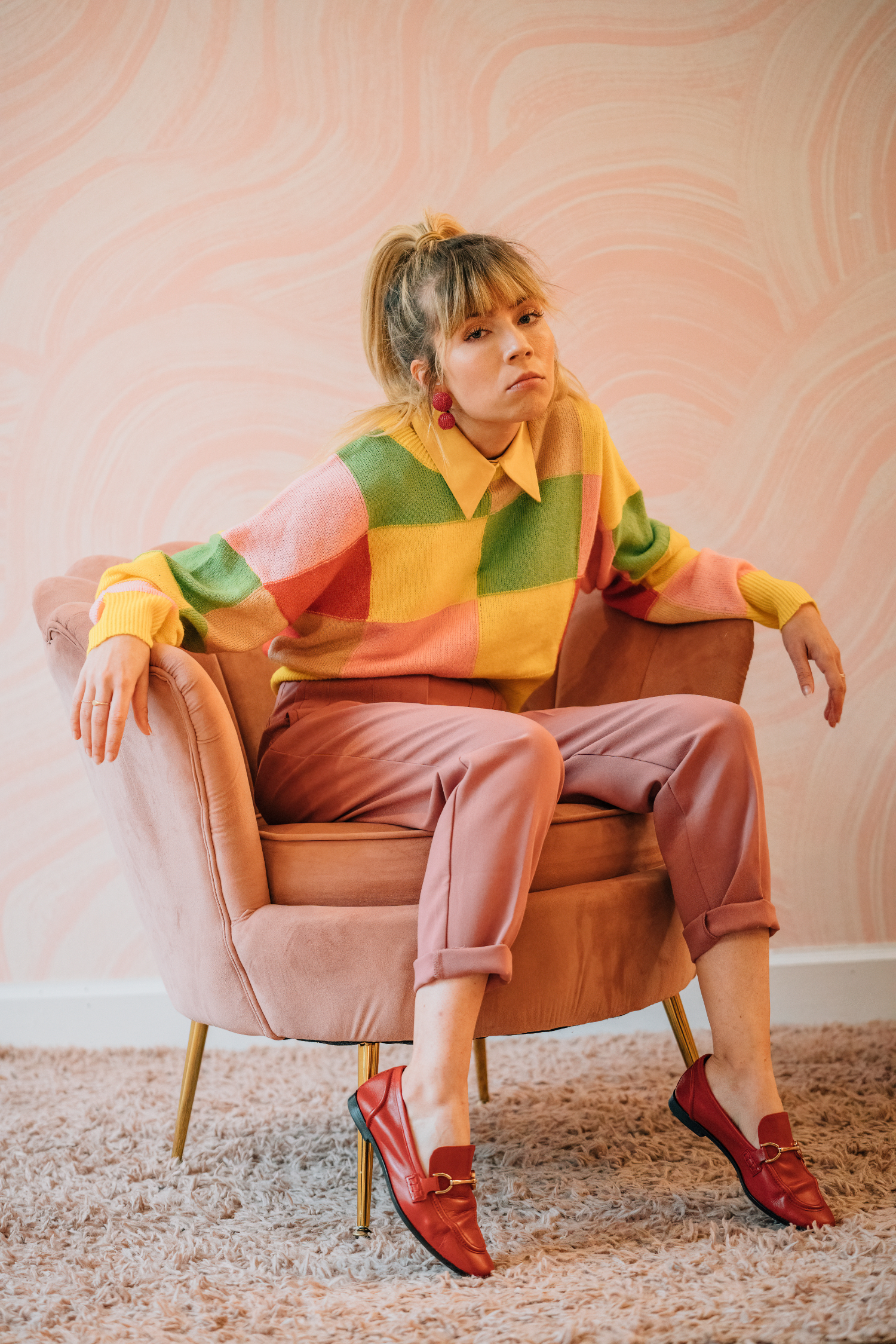 A person sits casually in a plush pink chair against a softly patterned pink wall. They wear a colorful patchwork sweater in shades of yellow, green, pink, and red over a collared shirt, paired with mauve pants and red loafers. Their expression is calm yet confident, and the warm tones of the outfit and setting create a playful, vintage-inspired aesthetic.