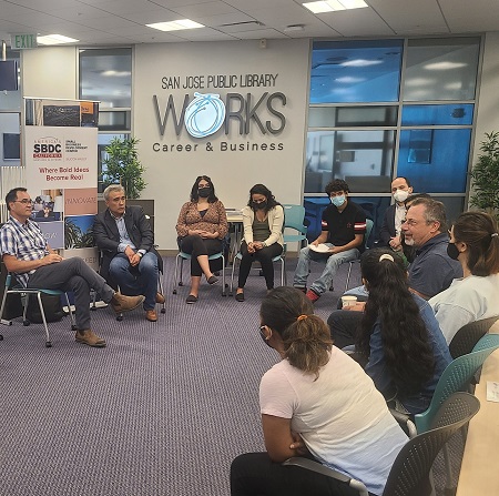 A group of male and female students sit in a circie, looking engaged. On the left a dark-haired man and an older gentleman appear to be speaking.