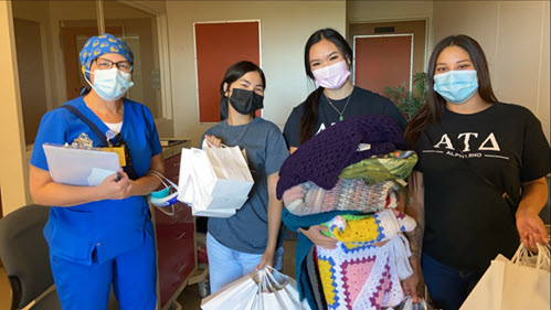 Alpha Tau Delta Volunteering 02 Three Alpha Tau Delta members donating items, including blankets. The three members are darked haired women, wearing masks while obviously smiling. A nurse, dressed in blue, stands and smiles with them.