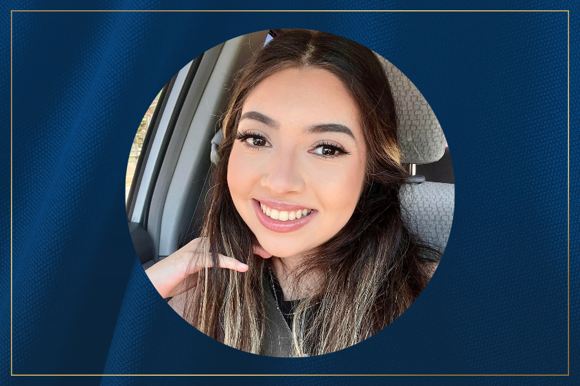 A person with long brown hair poses in a car, holding her hand up in a casual gesture.